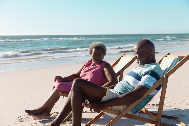 African american senior couple talking while relaxing on deckchairs against seascape and clear sky. Copy space, unaltered, vacation, love, together, retirement, summer, enjoyment and nature concept.