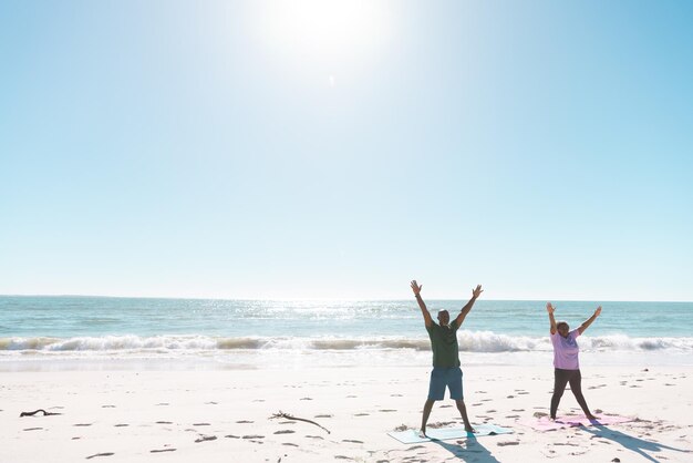 写真 海と澄んだ青い空を背景にビーチで腕を上げて運動するアフリカ系アメリカ人のシニアカップル。スペース、夏、変更されていない、愛、一緒、退職、自然、休暇、アクティブなライフスタイルをコピーします。