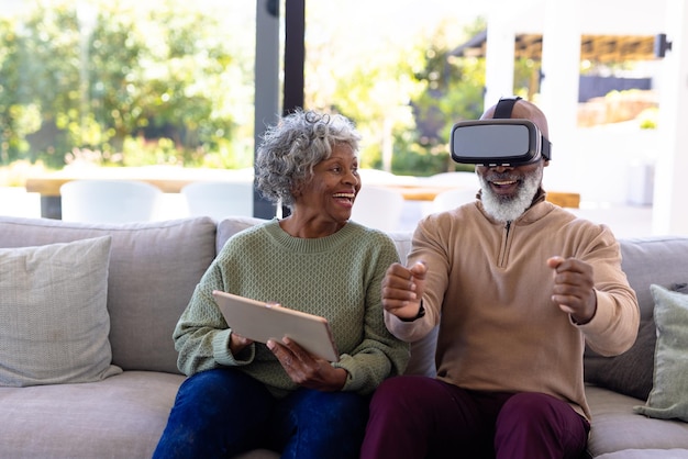 african american senior man enjoying virtual reality simulator and woman using digital pc on sofa. retirement home, unaltered, creative, gesturing, technology, together, support, assisted living.
