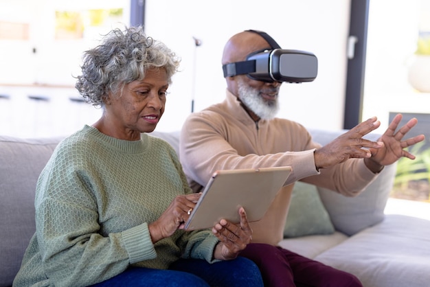 African american senior woman using digital pc and man using virtual reality simulator on sofa. Retirement home, unaltered, creative, gesturing, technology, togetherness, support, assisted living.