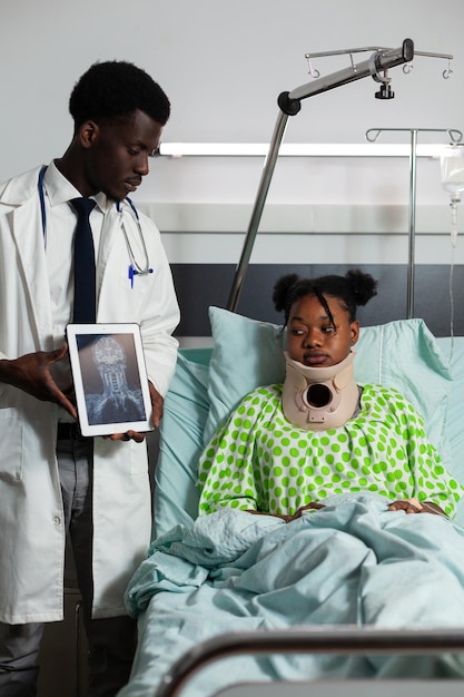 African american sick patient wearing neck cervical collar lying in bed recovering after medical surgery. Therapist doctor holding tablet with bones radiography on screen working in hospital ward