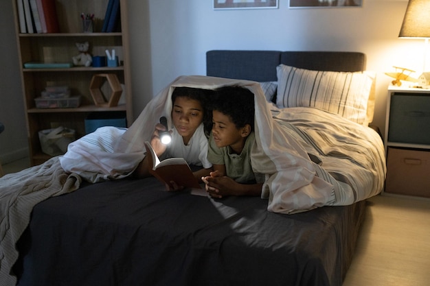 African american sister holding flashlight and reading book to brother