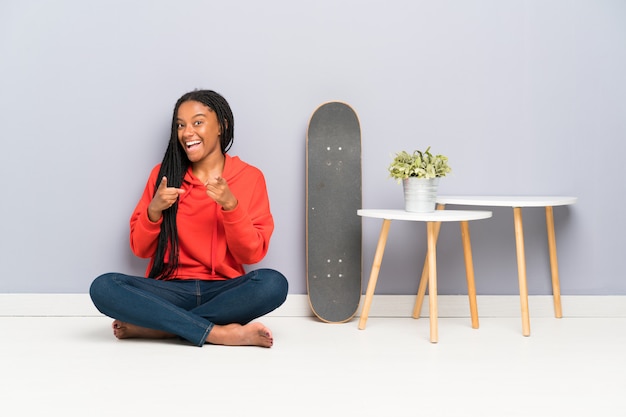 African American skater teenager girl with braided hair sitting on the floor points finger at you