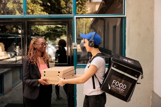 Foto impiegato sorridente afroamericano che riceve l'ordine della pizza all'aperto, donna che prende la pila di pacchetti pasto dal corriere. ragazza in cuffia che consegna pasti da asporto, servizio di consegna pizzeria