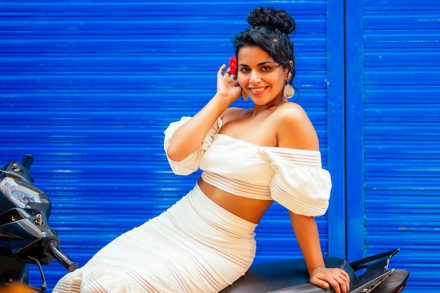 African american smiling woman in white dress posing on blue street wall on scooter