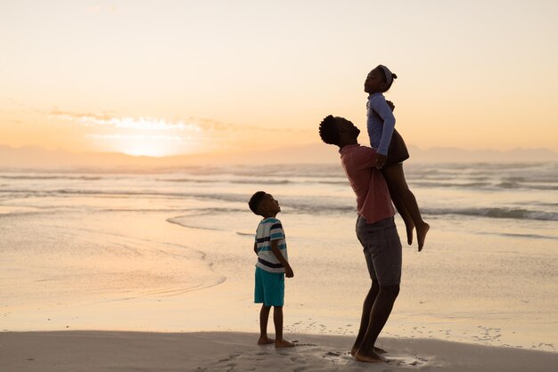 African american son looking at father picking up girl while standing on beach against sky at sunset