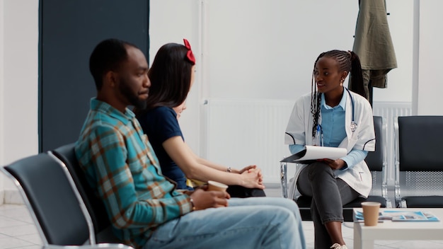 Афроамериканский специалист задает вопросы о здравоохранении, чтобы проконсультировать маленькую девочку с матерью в вестибюле зоны ожидания. Врач разговаривает с ребенком с мамой о болезни и диагнозе.