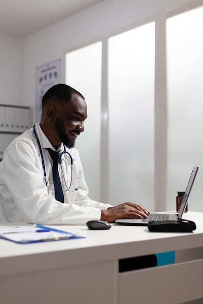 African american specialist doctor analyzing medicine prescription document report checking medical treatment on laptop computer. Therapist man working in hospital office. Healthcare service