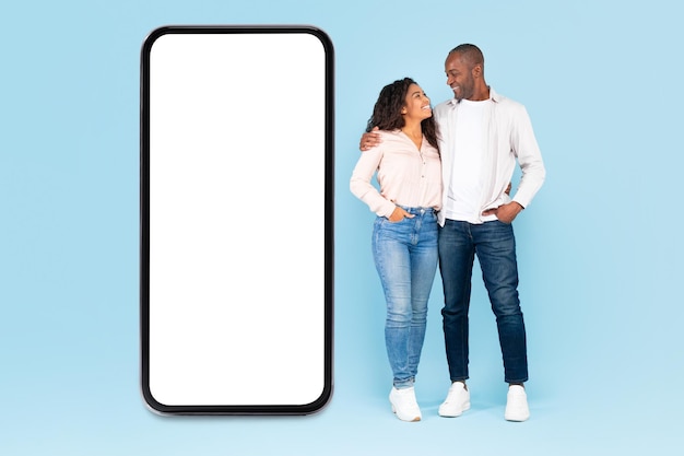 African american spouses standing near huge mobile phone with empty screen posing over blue background mockup