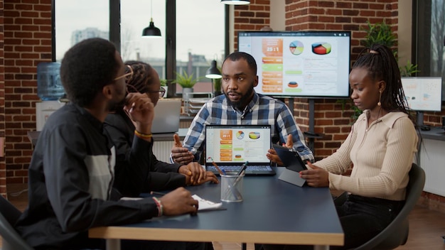 African american startup company brainstorming ideas to plan analysis with statistics on laptop and tablet. Office employees meeting to create financial charts and graphs, doing teamwork task.