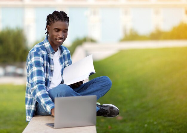 African american student guy websurfing on laptop holding workbooks outside