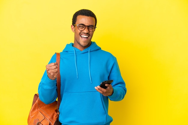 Афро-американский студент человек на изолированном желтом фоне с телефоном в победной позиции
