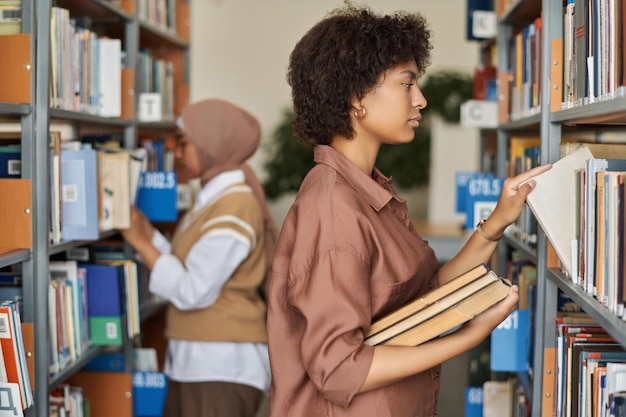 Афроамериканский студент стоит возле полок и ищет книги для изучения в библиотеке
