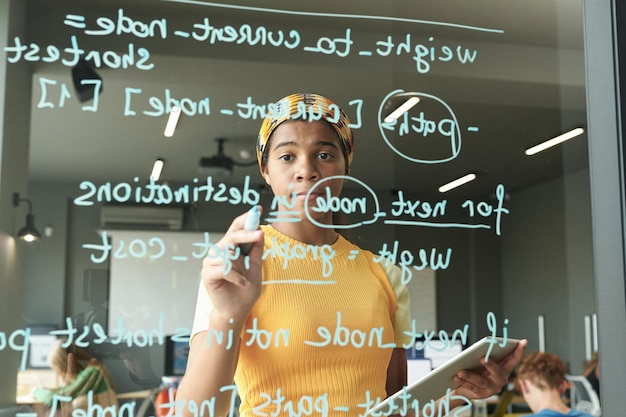 African American student using tablet pc and writing formulas on glass wall during lesson