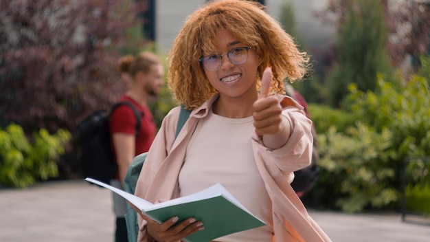 Афроамериканская студентка читает книгу на открытом воздухе, улыбается в камеру, показывая большой палец вверх в средней школе