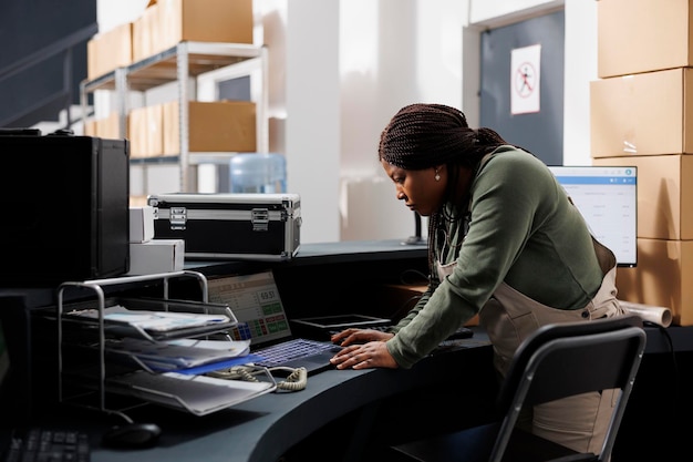 African american supervisor checking customers orders on laptop computer, analyzing shipping details in warehouse. Storage room employee wearing industrial overall working at products inventory report