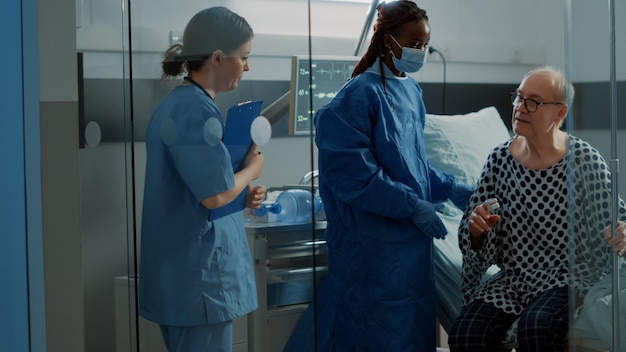 Photo african american surgeon walking elder patient from hospital ward with nurse at intensive care facility. sick old man connected to modern oximeter and iv drip bag leaving bed for surgery