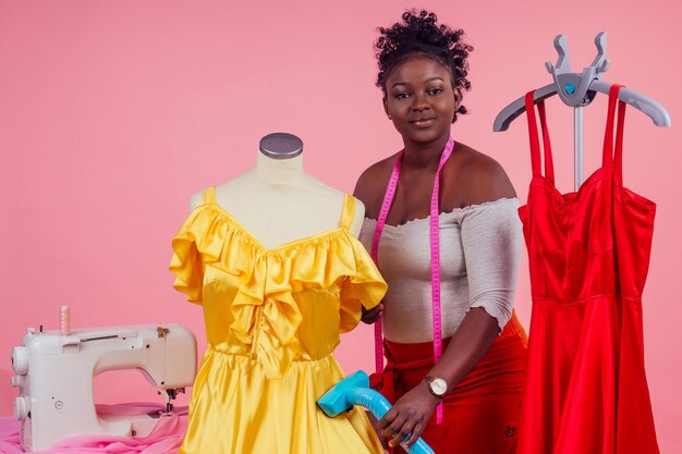 African american tailor dressmaker holding vapor device hot steam generator for silk red and yellow dress in dry-cleaning salon, studio pink background .