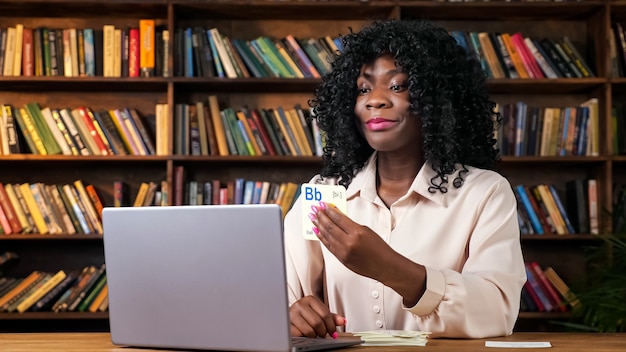 African-American teacher shows cards with letters to laptop camera at online English lesson sitting at table against bookshelves at home