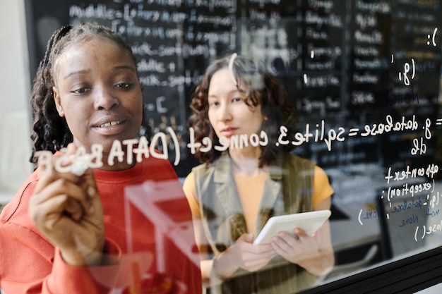 African american teacher writing codes on blackboard and explaining it to student in the classroom