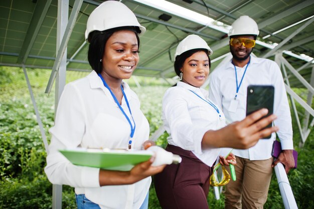 African american technician checks the maintenance of the solar panels. Group of three black engineers meeting at solar station. Make selfie by phone.