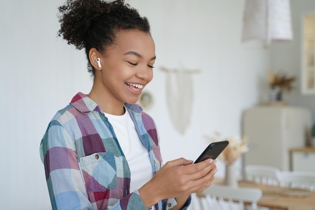 이어폰을 끼고 스마트폰을 들고 있는 아프리카계 미국인 10대 소녀는 집에서 음악 앱을 사용하여 오디오 메시지를 듣습니다.