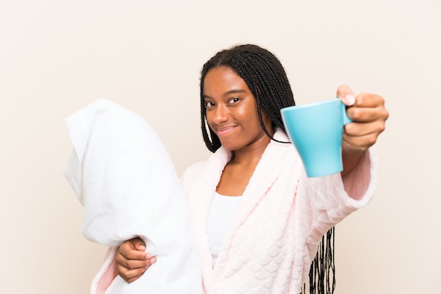 African american teenager girl with long braided hair in pajamas over wall and holding a cup of coffee