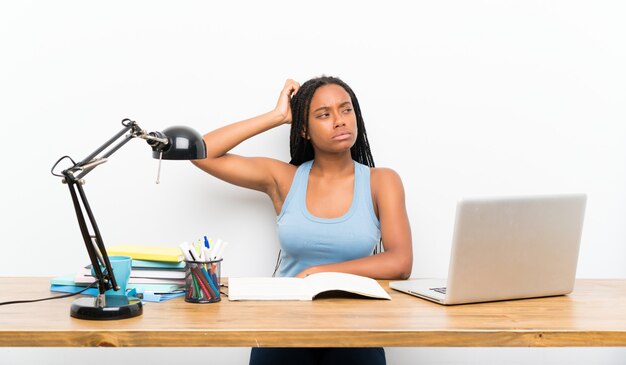 African American teenager student girl with long braided hair in her workplace having doubts while scratching head