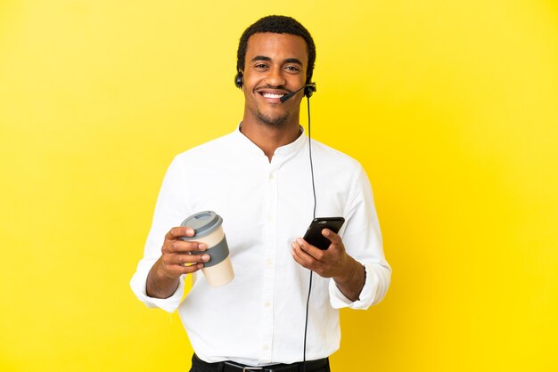 African american telemarketer uomo che lavora con un auricolare su sfondo giallo isolato che tiene il caffè da portare via e un cellulare