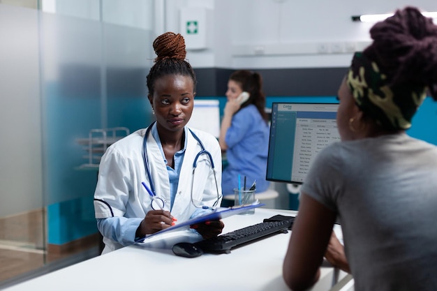 African american therapist discussing sickness symptoms questionnaire with sick patient during clinical appointment working in hospital office. Practitioner doctor explaining healthcare treatment