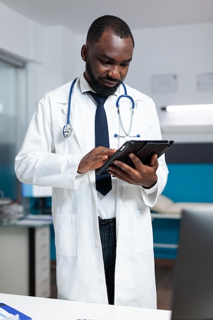 African american therapist doctor analyzing medicine prescription using tablet computer checking sickness expertise during medical appointment. Practitioner man working in hospital office