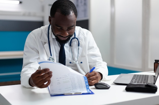 African american therapist doctor checking medicine prescription documents analyzing pharmaceutical treatment. Pactitioner man working at patient sickness report in hospital office