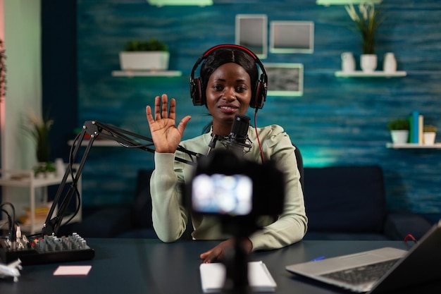 African american vlogger woman greeting subscribers while recording video content for social media. Influencer sitting in podcast studio filming tutorial using production equipment. On air talk show