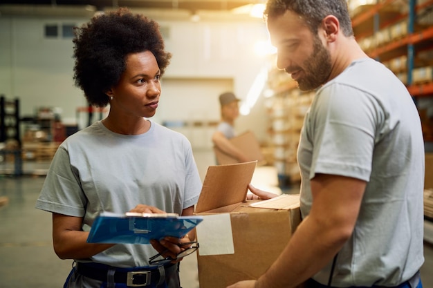 african american warehouse worker talking to her colleague while working at industrial storage compartment
