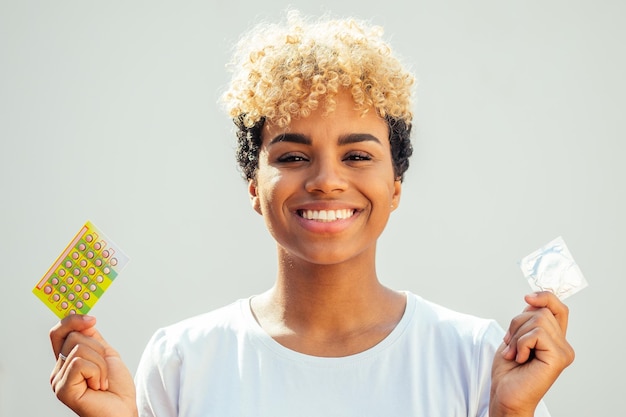 La donna afroamericana si perde nei suoi pensieri con una confezione di preservativo e pillole anticoncezionali scegliendo uno studio di sfondo bianco pensante