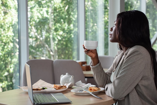 African american woman in business suit with laptop and pizza drinking tea in a cafe looking out the window.