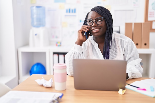 African american woman business worker using laptop talking on smartphone at office