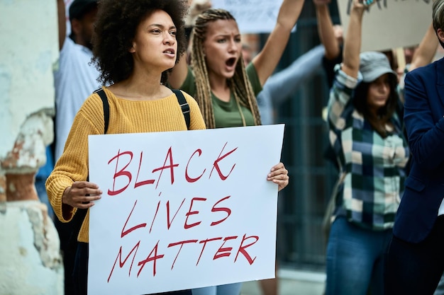 路上で人々の群衆に抗議するブラック・ライヴズ・マターのバナーを運ぶアフリカ系アメリカ人の女性