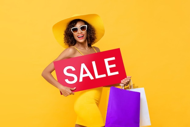 스튜디오 노란색 배경에 격리된 쇼핑백과 함께 빨간색 판매 표지판을 들고 있는 아프리카계 미국인 여성