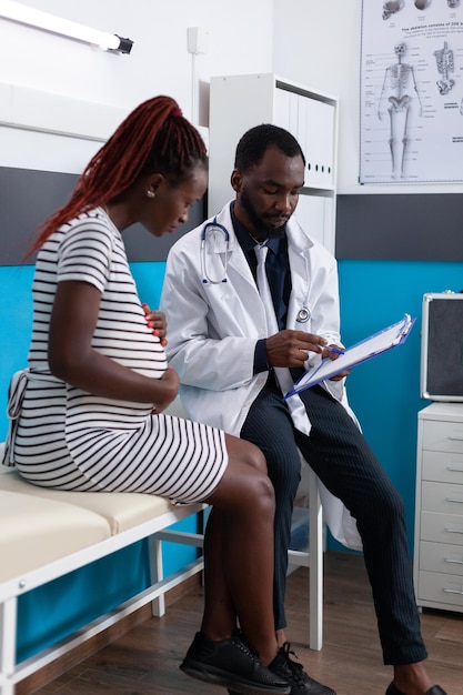 Donna e medico afroamericani che parlano di gravidanza all'appuntamento nel gabinetto. paziente in attesa di un bambino che si consulta con il medico per controllare l'assistenza sanitaria. incinta adulta