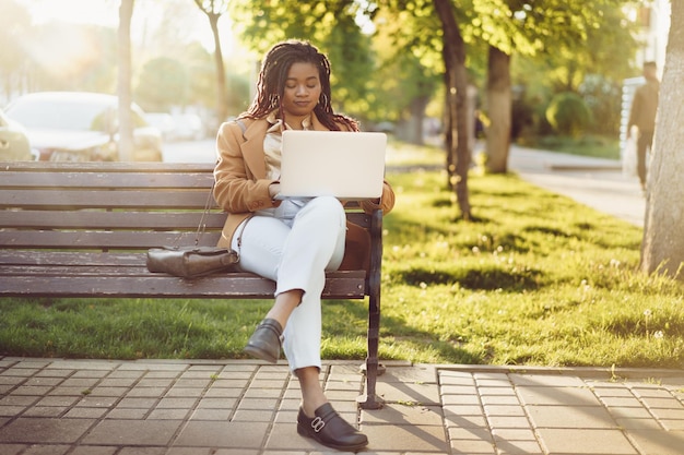 African american woman freelancer sitting on a bench in a street and using laptop