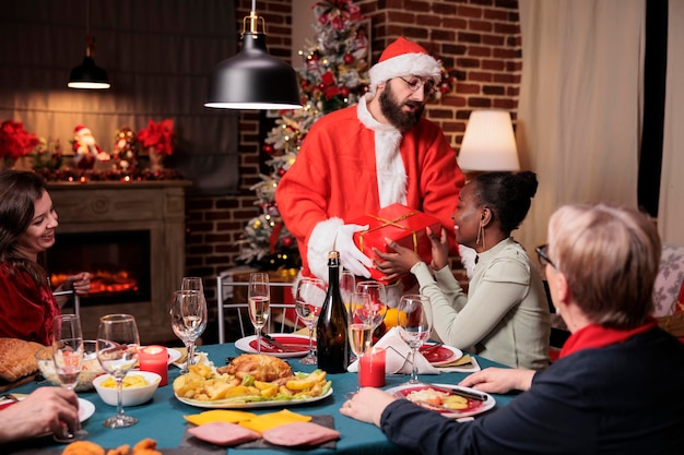 African american woman getting gift from man in santa claus\
costume, family exchanging christmas presents. friends celebrating\
xmas eve, eating traditional meal at festive dinner table