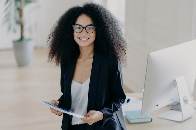 African American woman holds some papers, wears elegant clothes and spectacles