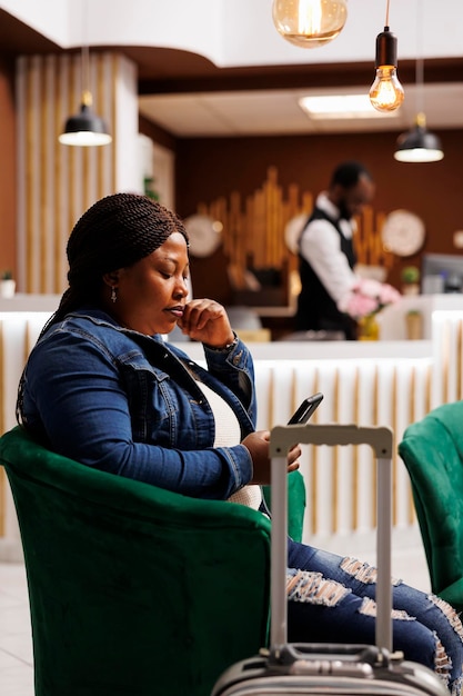 African american woman hotel guest using mobile phone, sitting in armchair with smartphone waiting for check-in. black female tourist sits in lobby with suitcase ordering taxi via mobile phone app