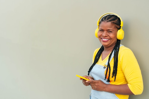 Афро-американская женщина слушает музыку и использует смартфон, стоя на сером фоне.
