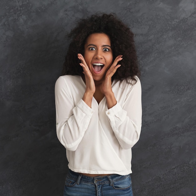 African-american woman looking excited, surprised in full disbelief, touching face on black studio background