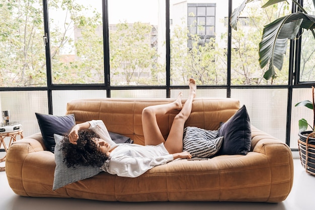 African american woman lying down on sofa with legs up in a bright loft living room Home concept