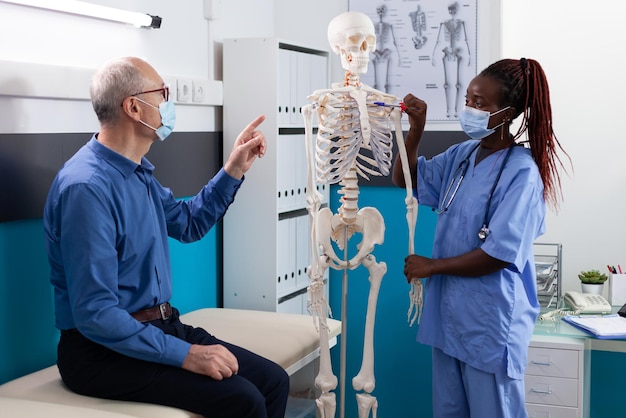 African american woman nurse with medical protective face mask against covid19 holding human skeleton model explaining spine pain to senior man patient in hospital office. Osteoporosis concept