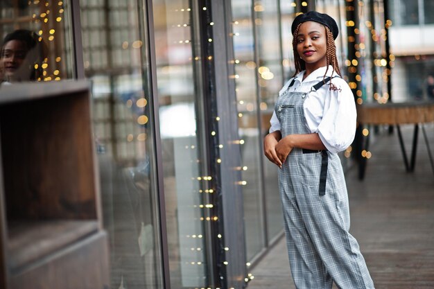 African american woman in overalls and beret posed in outdoor terrace with christmas decorations garland