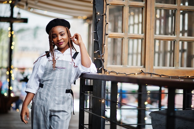 African american woman in overalls and beret posed in outdoor terrace with christmas decorations garland.